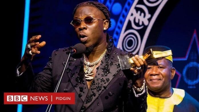 cdd05db0-2097-11ef-a2c1-6ff4d291c70e.jpg A man wearing sunglasses and a black outfit speaks into a microphone while holding an award, with another man smiling beside him on stage. BBC News Pidgin logo is visible, highlighting Stonebwoy among TGMA 2024 winners for artiste of the year.