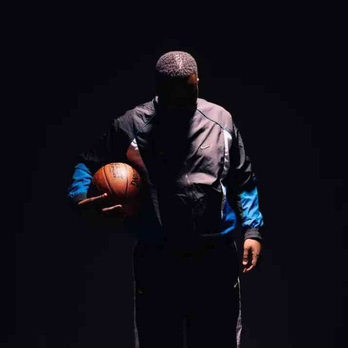 img_1_1744960009448.jpg A person in a dark tracksuit stands holding a basketball under one arm, partially illuminated against a dark background, channeling the cool confidence of Sfarzo Rtee.