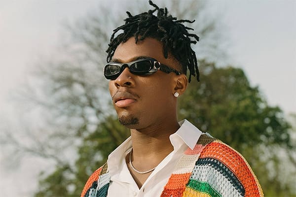 joeboy.jpg Person with sunglasses and a colorful sweater stands outdoors with trees in the background, channeling Joeboy's vibrant Afrobeats vibe.