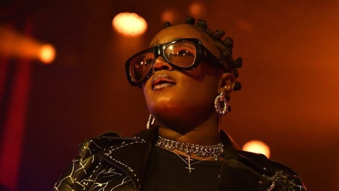 media_1a02e2ab897c03ab1a7620fed32477aa97d19ed56.jpeg A female African artist, reminiscent of FAVE or Tems, stands under warm stage lighting. She's wearing large glasses and a leather jacket, with studded earrings and a necklace, her gaze directed upwards.
