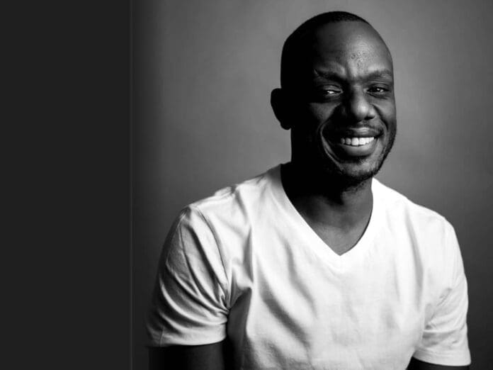 theglobalriseofamapiano.jpg Black and white portrait of a man in a white t-shirt smiling at the camera against a plain background, capturing the spirit of SA Music and the global rise of Amapiano.