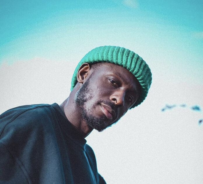 53768736.jpg A man wearing a green knit beanie and a dark shirt looks at the camera against a bright blue sky, embodying Kaas The Messenger’s Afrobeats Gospel vibe.