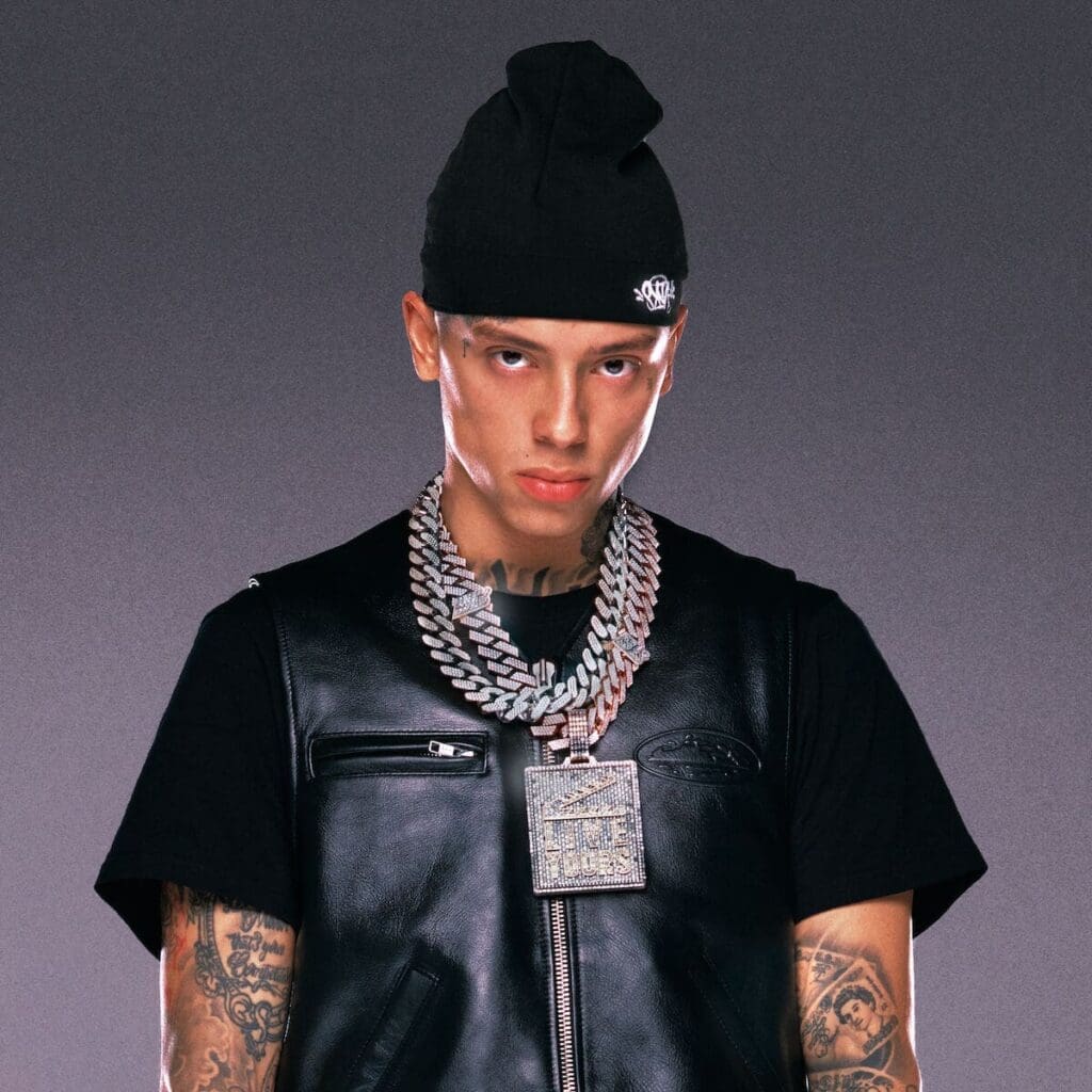A man wearing a black beanie, black t-shirt, black leather vest, and multiple large silver chains stands against a plain gray background.