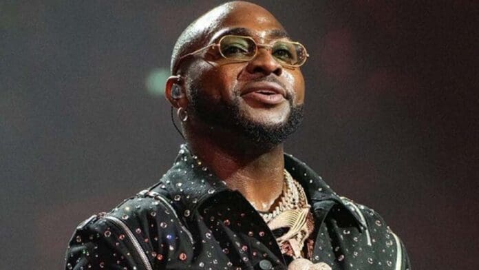 DAVIDO-1-1.jpg A man wearing glasses, a jeweled black shirt, and layered necklaces stands under stage lighting, channeling Afrobeats icons like D’banj and Davido as he looks forward.