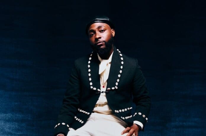 DAVIDO-press-credit-Chisean-Rose-2025-billboard-1548.jpg A man wearing a black studded jacket, white shirt, and black leather cap sits against a dark background, looking at the camera with a neutral expression—much like an artist ready for his Billboard debut on the U.S. Afrobeats Songs Chart.