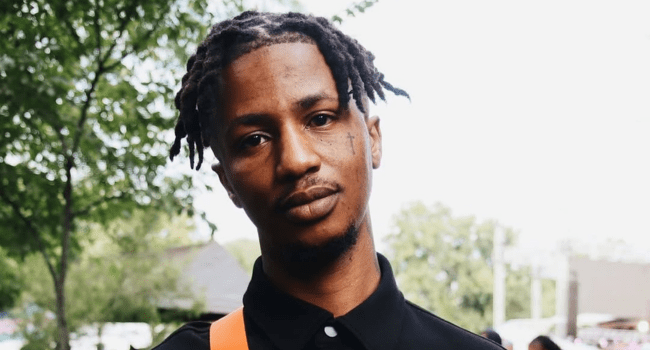 Emtee-11.png A man with short, twisted dreadlocks and a small face tattoo stands outdoors wearing a black collared shirt with an orange strap—Emtee, recognizable to fans and Metro FM Award winner. Trees and sky form the background.
