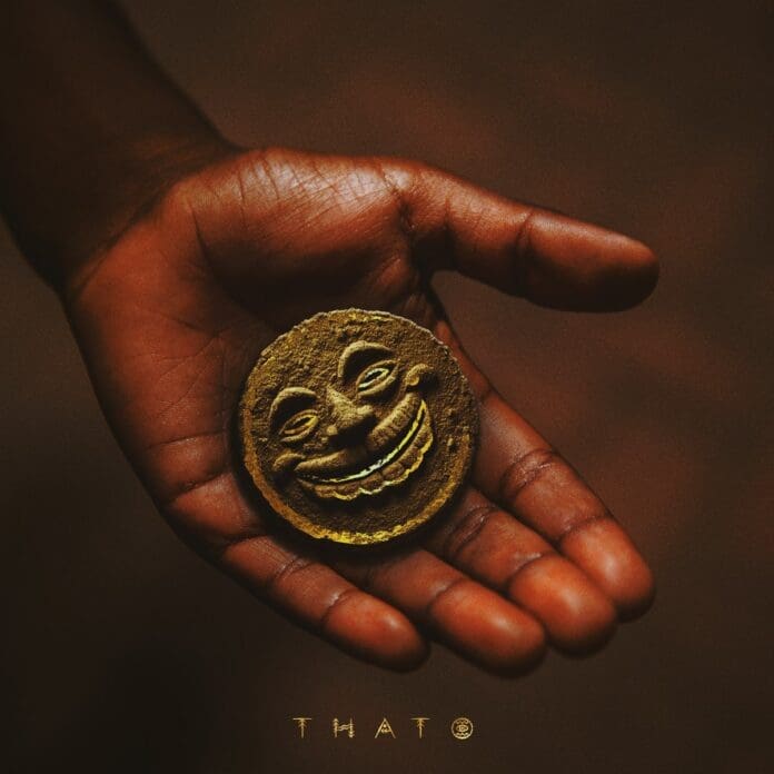 Kelvin-Momo-–-Thato-Ya-Modimo-Album-4.jpg A hand holds a round, gold-colored object with a carved smiling face, reminiscent of the vibrant energy in Kelvin Momo and Da Muziqal Chef’s Emhlabeni.