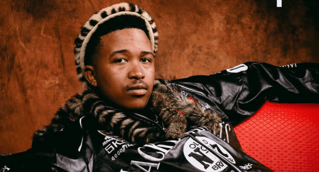 Lowfeye-5.png A man wearing a black jacket with white lettering and traditional patterned accessories sits on a red seat against a brown background—perfect for a Spotify Cover or highlighting his New Single Release.