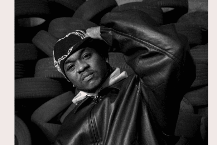f2314db6-ee2a-4113-97d3-aa686ffa15ef.png A person wearing a beanie and leather jacket poses in front of a stack of tires, channeling Afropop vibes and looking directly at the camera with a neutral expression.