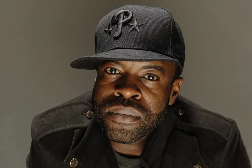 The Roots with a beard wearing a black jacket and a black baseball cap with the letter "P" and two stars, looking directly at the camera against a neutral background.