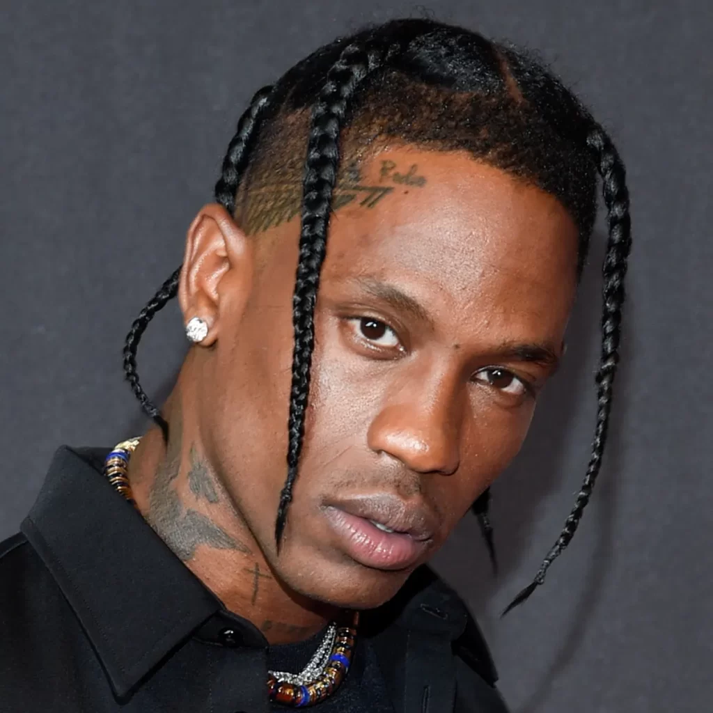 Travis Scott with braided hair, facial tattoos, diamond earrings, and a black shirt looks toward the camera against a dark background.