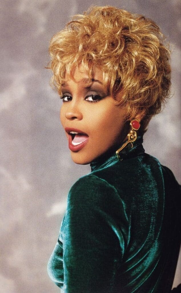 Whitney Houston
with short, curly blonde hair wearing a green velvet top and bold earrings poses with her mouth slightly open against a mottled gray background.