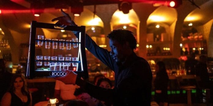 a-waiter-brings-drinks-to-customers-at-a-restaurant-lounge-club-in-lagos.jpg A server holds a tray of shot glasses in a dimly lit bar with red and yellow lighting, Afrobeats playing softly and patrons seated at tables in the background, capturing the vibrant spirit of Lagos nightlife.