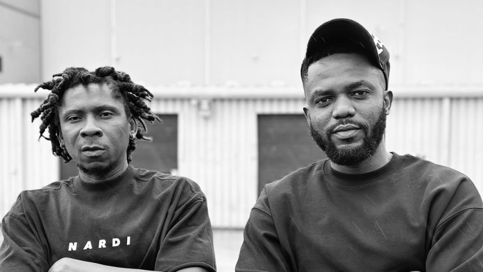 Two men stand side by side outdoors in front of a corrugated metal building, both in plain dark shirts; one wears a cap and the other has dreadlocks. The black and white image channels the spirit of Ghanaian classics.