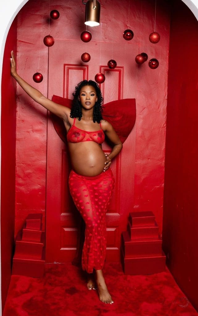 1000000242.jpg A pregnant woman in a sheer red outfit poses barefoot in a red alcove decorated with hanging red ornaments, capturing an elegant moment reminiscent of Amapiano vibes and Central News South Africa style.