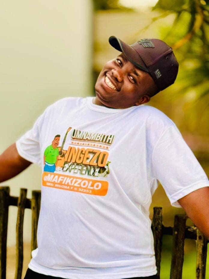 A man wearing a black cap and a white graphic t-shirt stands outdoors, smiling broadly with arms resting on a wooden fence, showing his pride in Mzansi Artists who made headlines in Central News South Africa.