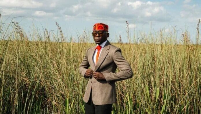 A person wearing a tan blazer, orange tie, sunglasses, and a red patterned headwrap stands in a grassy field under a partly cloudy sky, channeling SPINALL’s Èkó Groove vibes with an unmistakable Afrobeats flair.