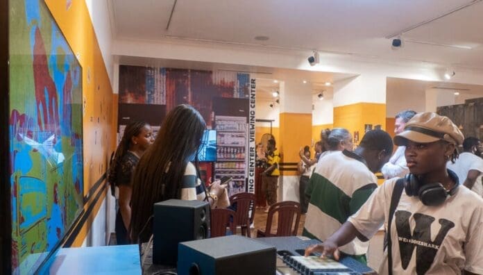 People interact and observe displays at an indoor art exhibition, with Afrobeats music equipment visible in the foreground and colorful artwork on the walls—a vibrant scene often highlighted by OkayAfrica.