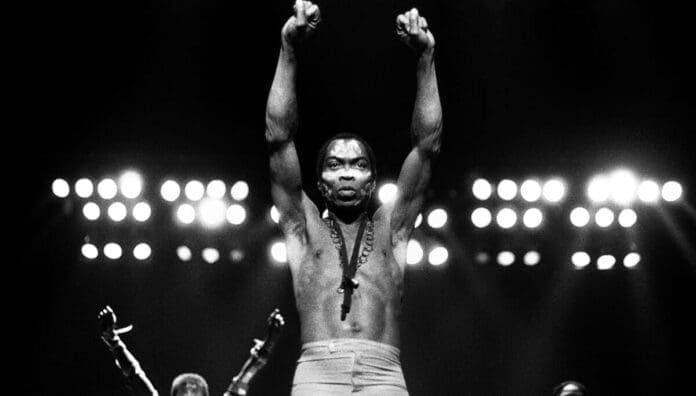 A shirtless man stands on stage with both arms raised, fists clenched, under bright lights—reminiscent of Fela Kuti accepting a Lifetime Achievement Award at the Grammys.