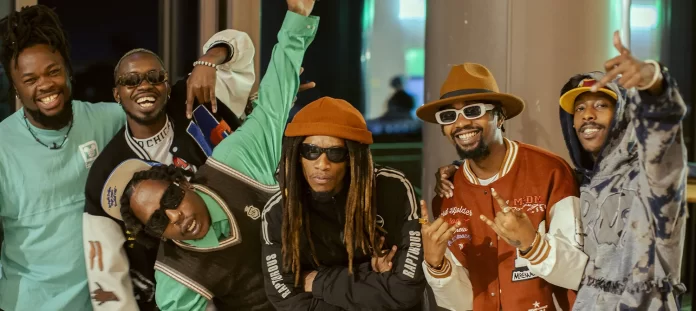 Six men pose together indoors, smiling and making gestures; they wear colorful streetwear inspired by Afrobeats culture, including hats, sunglasses, and layered clothing.