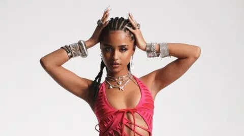 Woman wearing a red cut-out top, silver jewelry, and braided hair—channeling Tyla's influence on the African music market—poses with hands on her head against a plain light background.