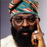 A man with a full beard and glasses, featured at 3Music, wears a colorful patterned headwrap and light clothing, holding his hand to his face and displaying rings—truly embodying "They’ll Call Us Crazy Before They Understand Us.