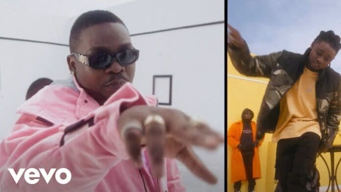 Two men in stylish outfits gesture to the camera in split-screen; one, resembling Olamide, wears a pink jacket and sunglasses, while the other sports black and green clothing. Vevo logo visible.