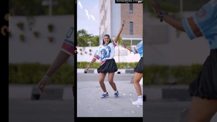 Two people wearing sports jerseys and skirts dance outdoors on a pavement, one mid-motion and smiling, showing off their moves for the Chella dance challenge. A building and greenery are visible in the background.