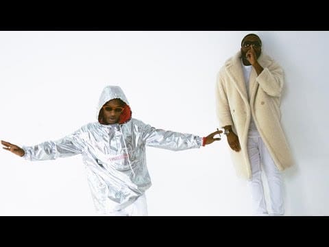 Two men pose against a plain white background; one, channeling Wizkid style, wears a shiny silver hooded jacket with arms outstretched, while the other sports a long cream-colored fur coat and sunglasses.