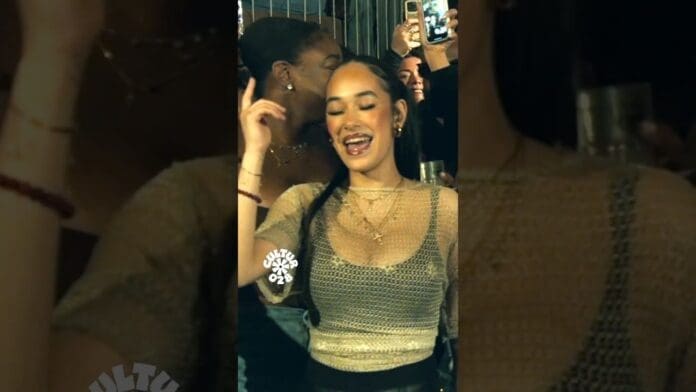 A woman smiles and dances at a party to an afrobeats DJ, surrounded by others. One person in the background is holding up a phone to record or take a photo.