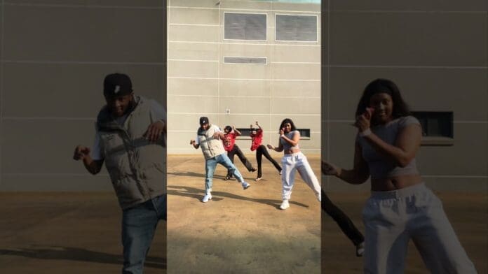 Four people groove energetically outdoors in casual clothing, performing an Amapiano Dance with a plain building wall in the background.