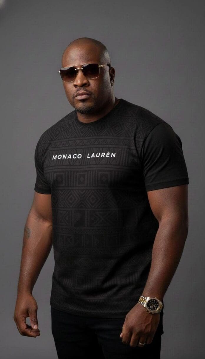 A man wearing sunglasses and a black "Monaco Laurèn" t-shirt stands against a plain grey background, channeling the fresh vibe of the Durban music scene.