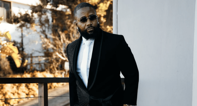 A man with a beard wearing sunglasses, a formal black suit, and a white shirt stands outdoors near a balcony railing, reminiscent of the stylish vibe at events like Fill Up Toyota Stadium featuring artists such as Cassper.