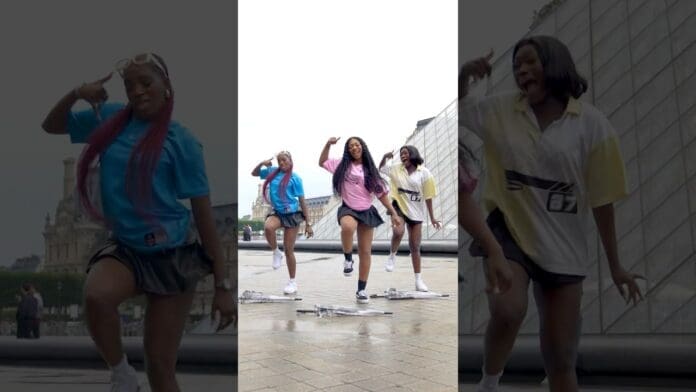 Three women in colorful outfits dance energetically outdoors on a paved area, performing lively choreography to Rema’s AZAMAN. A modern glass building and historic architecture provide a striking backdrop for this vibrant dance video.