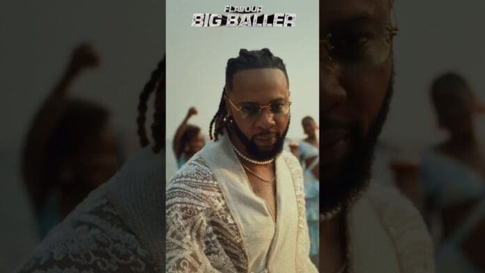 A man wearing sunglasses, jewelry, and a lace shirt poses in front of people with raised arms, exuding the vibe of African royalty. The text "Flavour Big Baller OFFICIAL VIDEO" appears at the top of the image.