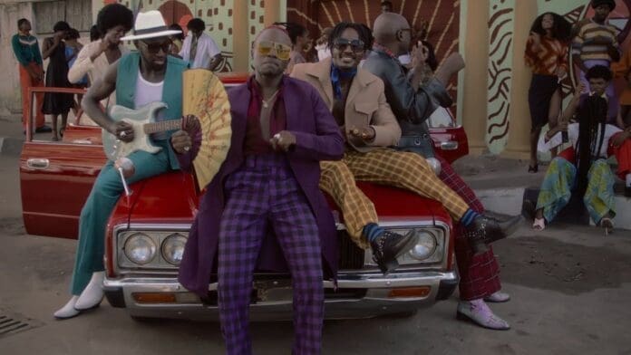 Four men in colorful, retro-style outfits sit on the hood of a red convertible, evoking vibrant city vibes—just like in the Sauti Sol "Suzanna" Official Video. Others in similar attire mingle in the lively street scene behind them.