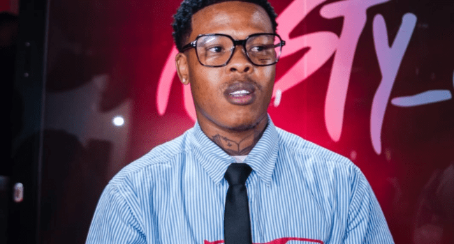 A person wearing glasses, a striped shirt, and a black tie stands in front of a pink and black background with stylized text, capturing the bold vibe of the star-studded line-up at the Durban Ivyson Tour featuring Nasty C.
