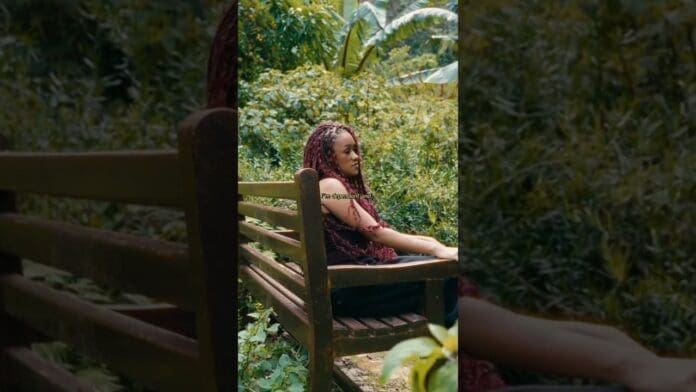 A person with braided hair sits on a wooden bench outdoors, surrounded by lush green plants and trees, quietly listening to new music—perhaps the latest RnB and Afrobeats hits.