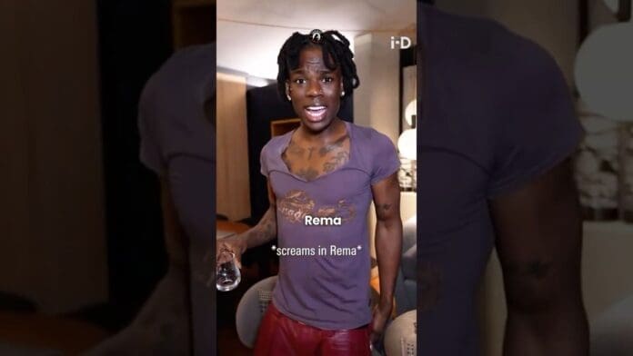 A person with tattoos, wearing a purple shirt and red pants, stands indoors holding a glass. Text on the image reads "Rema *screams in Rema*" as if he's ready to roast his fans at the show.