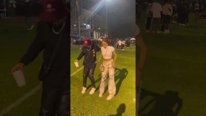Two people holding drinks walk on a grassy field at night, surrounded by other people and event tents, enjoying the vibrant energy of an Amapiano 2023 party.