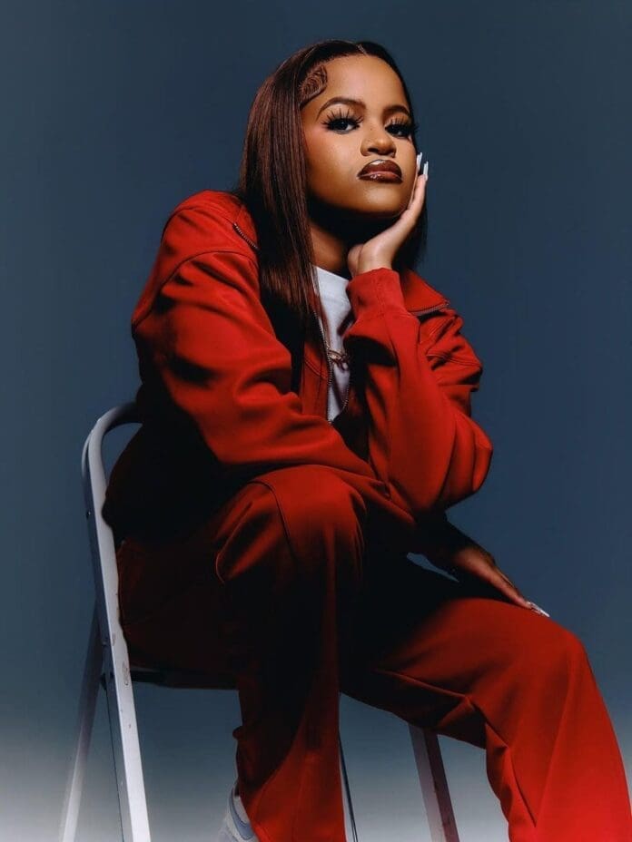 A person wearing a red tracksuit sits on a white folding chair, resting their chin on their hand, looking confidently at the camera—capturing the bold style of amapiano artist JayJayy against a dark background.