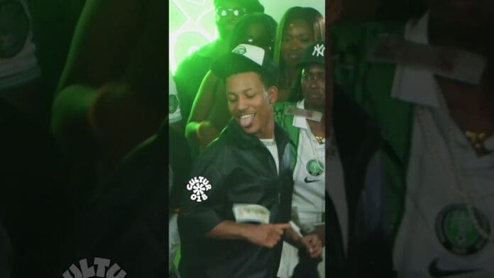 A man in a black jacket and cap dances with his tongue out, while dollar bills fly through the air. People in sports jerseys and hats watch from the background—looks like an energetic afrobeatmix party.
