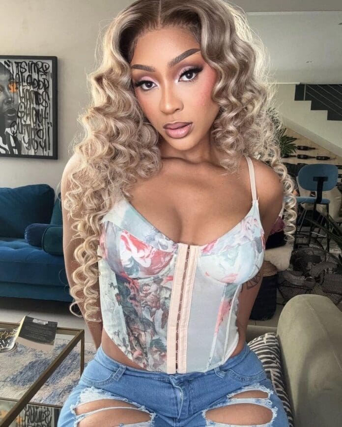 A person with long, curly blonde hair poses indoors, channeling Amapiano vibes in a floral corset top and ripped blue jeans. Modern furniture and artwork provide a stylish backdrop.