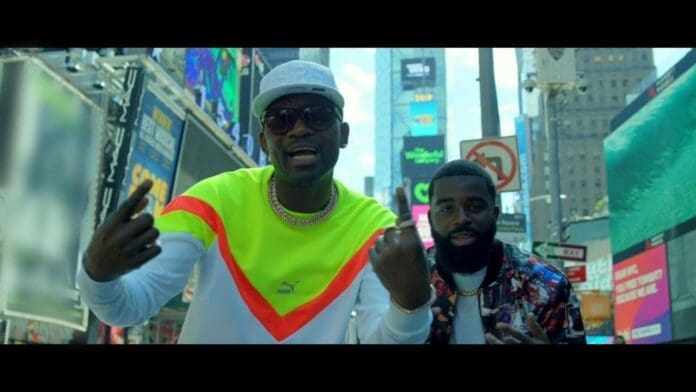 Two men stand in a busy urban area with bright digital billboards behind them; one wears a neon yellow and white sweatshirt and cap, the other has a beard and floral shirt, channeling vibes from an Official Music Video by Afro B and Busy Signal.