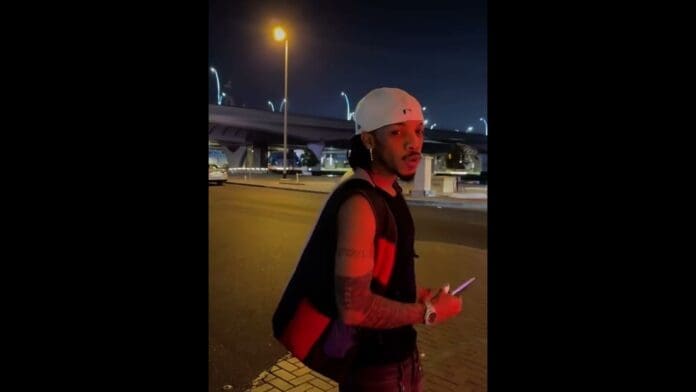 A man wearing a white cap, sleeveless shirt, and holding a phone stands on a city street at night, possibly catching a preview of Tekno's new song, with a bridge and streetlights in the background.