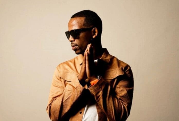A man wearing sunglasses and a brown jacket stands against a plain background with his hands together, looking to the side—reminiscent of Zakes Bantwini at the South African Music Awards, making waves in the music industry.