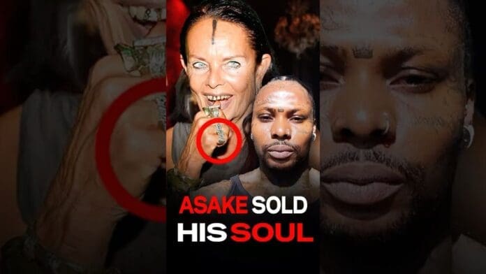 A woman displays a ring with a green stone while baring her teeth; beside her is a man. The text reads, "Asake sold his soul," hinting at ties to a secret society or Hollywood intrigue.