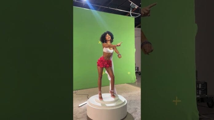 A woman in a white top and red skirt stands on a round platform, posing and pointing, in front of a green screen studio—perfect for a YouTube shoot or promoting the freshoutthegallery ALBUM OUT NOW.