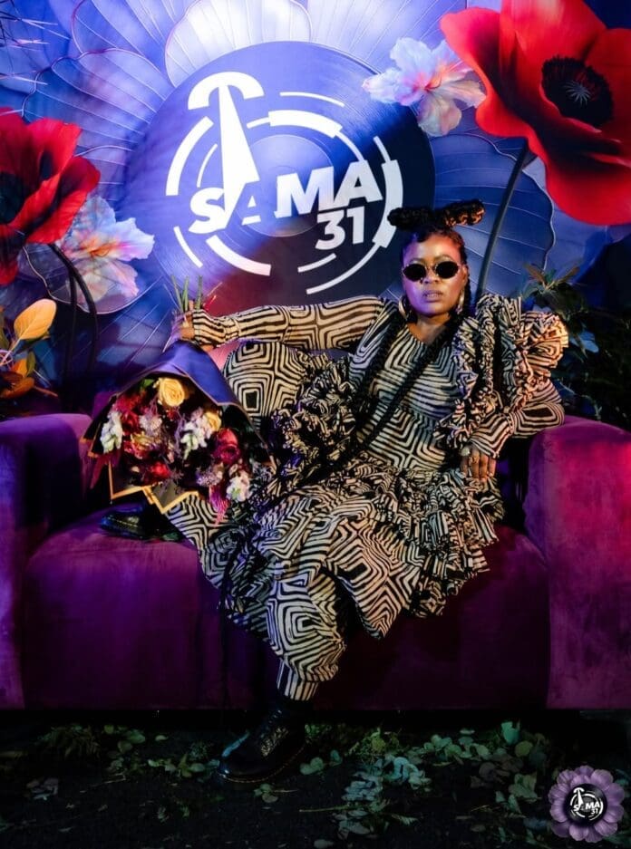 A person in patterned clothing and sunglasses sits on a purple couch with a bouquet, against a vibrant floral backdrop featuring the South African Music Awards SAMAs 31 logo.
