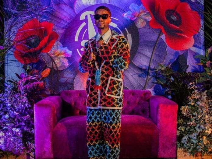 A person wearing a colorful patterned suit and sunglasses stands in front of a purple sofa, surrounded by vibrant flowers and a decorative backdrop, bringing true Mzansi flair to the scene—perfect for thestar.co.za’s 2025 Samas coverage.